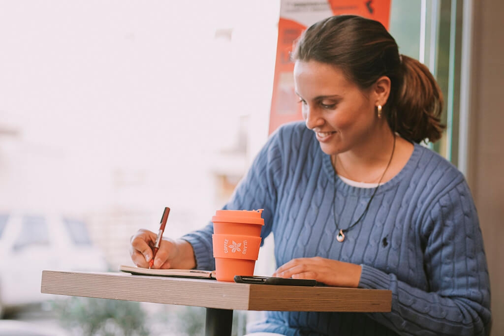 A woman writing in her notepad while enjoying her tea in a reusable cup from Coffee Island
