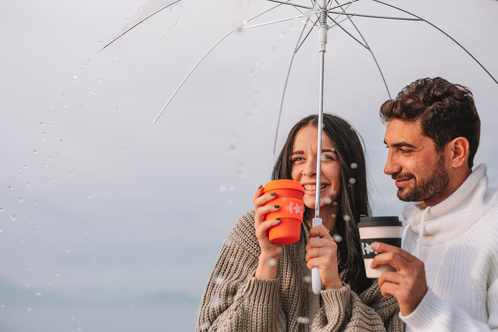 A man and a woman under their umbrella holding their coffee island reusable cups. 