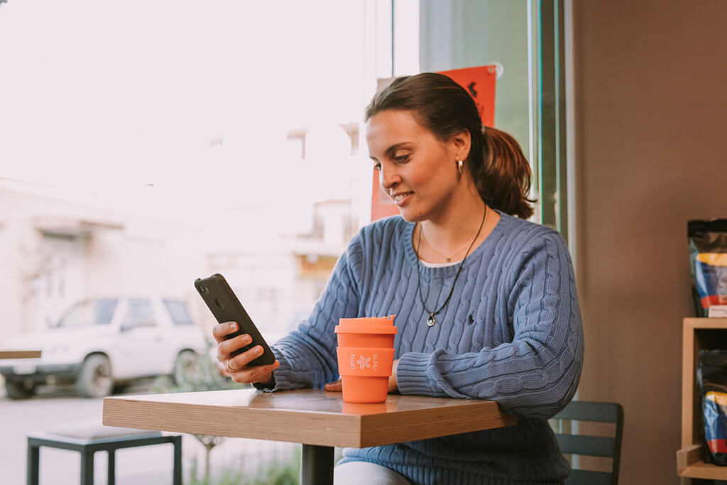 A woman sitting at a Coffee Island store, while enjoying her coffee with a reusable cup.